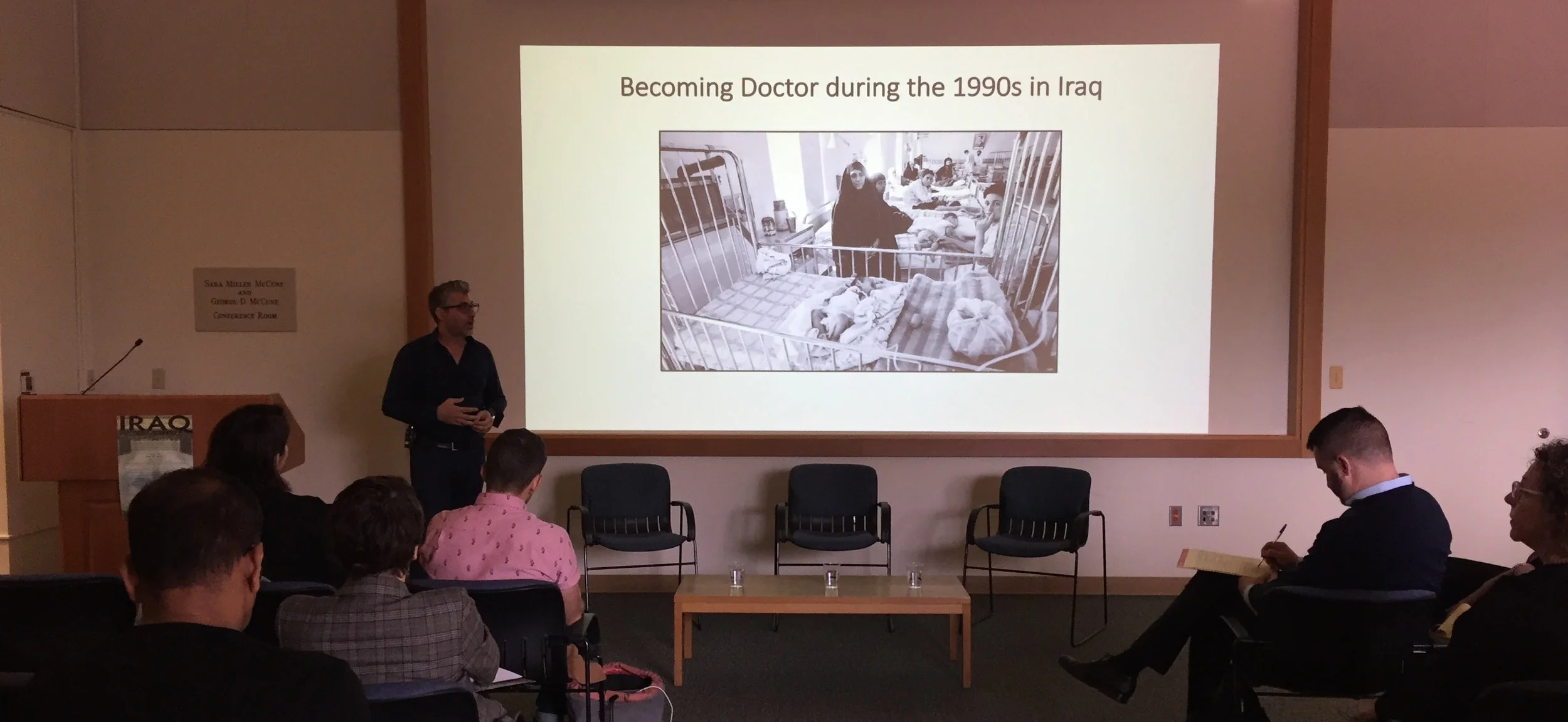  Dr. Omar Dewachi (next to screen) presents his lecture, titled “Iraqibacter: Ecologies of War and an Anthropology of Wounding” on the second day of the Iraq Front and Center symposium. 
